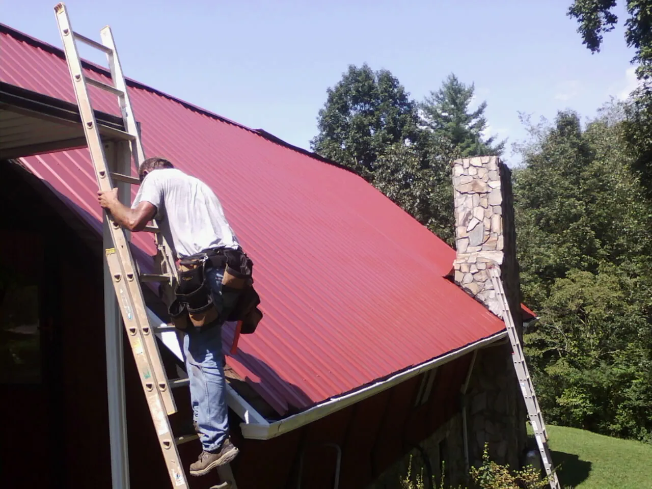 Roofer on a ladder beside a red metal roof and stone chimney — Soffit & Fascia Repair by Roofing Brothers in Boston